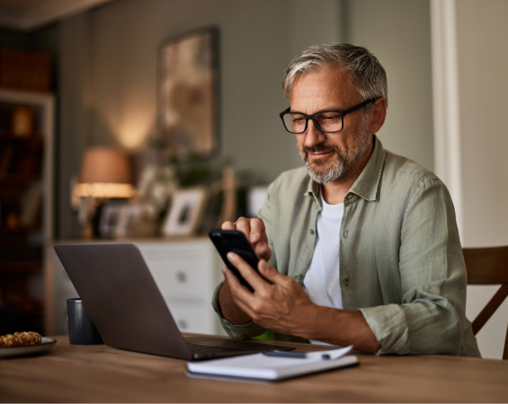 Middle Aged Teacher, Sitting At His Dining Room Table At Home Trying Out His New Smartphone.