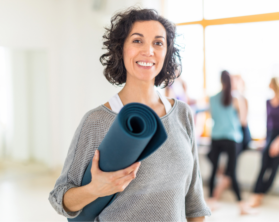 Middle aged female teacher at a yoga class, holding her mat, about to go in for a discounted class.