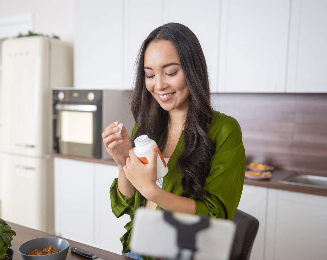 A young teacher opening a vitamin supplement bottle and reading the description while she preps dinner.